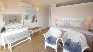 A hotel room with a bed, two white wicker chairs, a white desk with a bench, a television, and a wall-mounted air conditioner. - Fairlight Beach House (eMdloti)