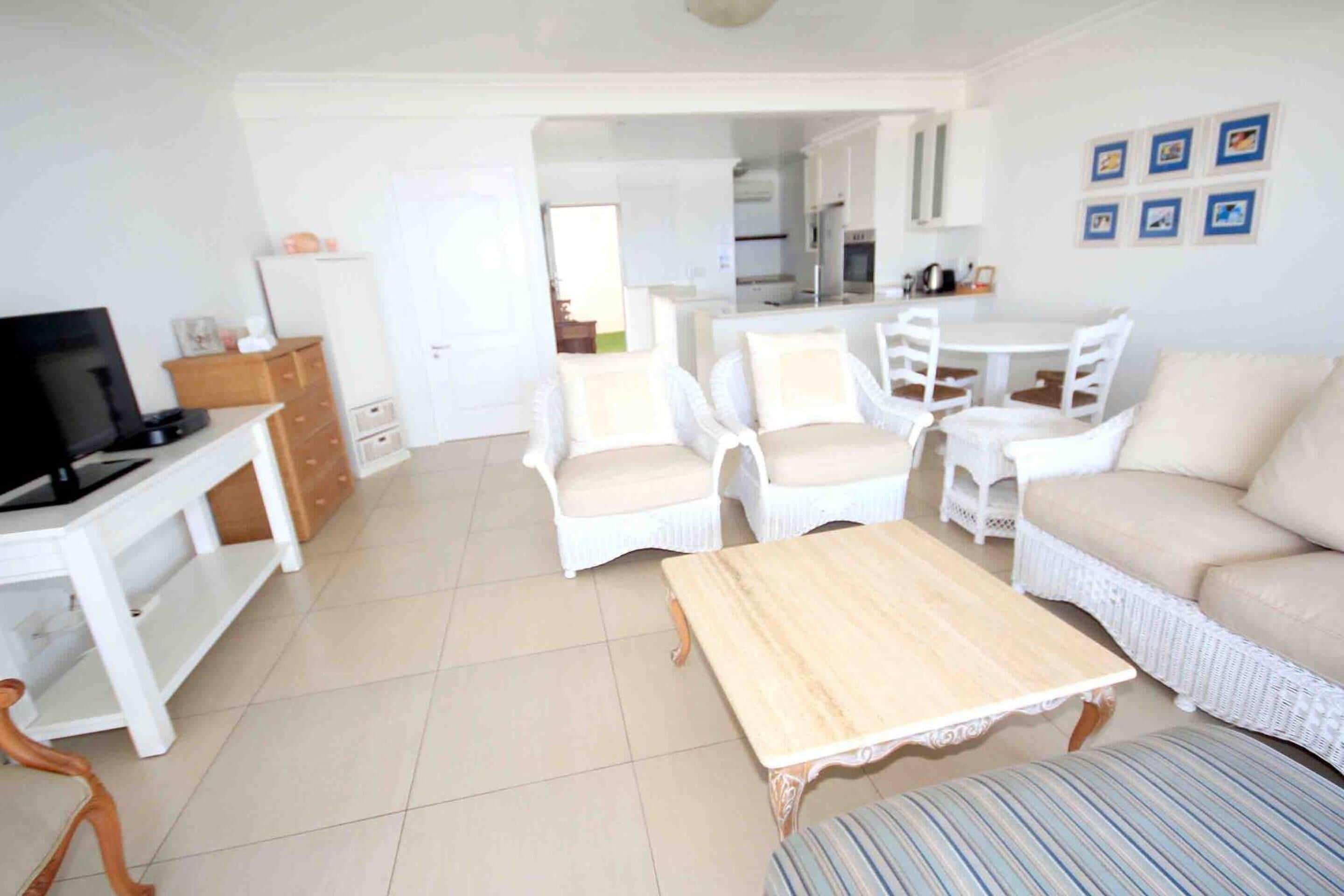 Un salón amplio con televisión, muebles de mimbre blanco, una mesa de centro de madera y un comedor con una mesa y sillas blancas.