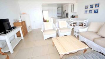 Un salón amplio con televisión, muebles de mimbre blanco, una mesa de centro de madera y un comedor con una mesa y sillas blancas.
