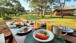 Un área de comedor al aire libre con una mesa preparada para el desayuno, incluyendo platos de comida, vasos de jugo y una taza.