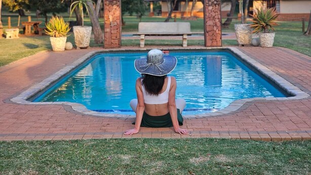 Una persona sentada junto a una piscina rectangular con borde de piedra, rodeada de plantas en macetas y un banco.