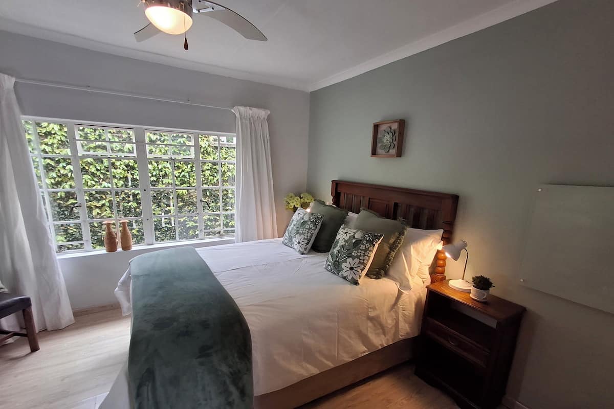 A bedroom with a large bed, a nightstand with a lamp, a window with curtains, and a ceiling fan.