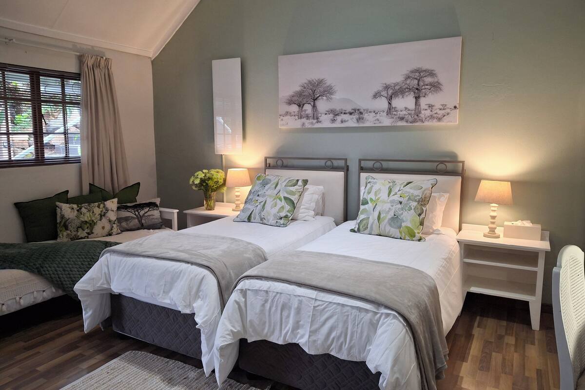 A bedroom with two beds, a window with curtains, a painting on the wall, and bedside lamps.