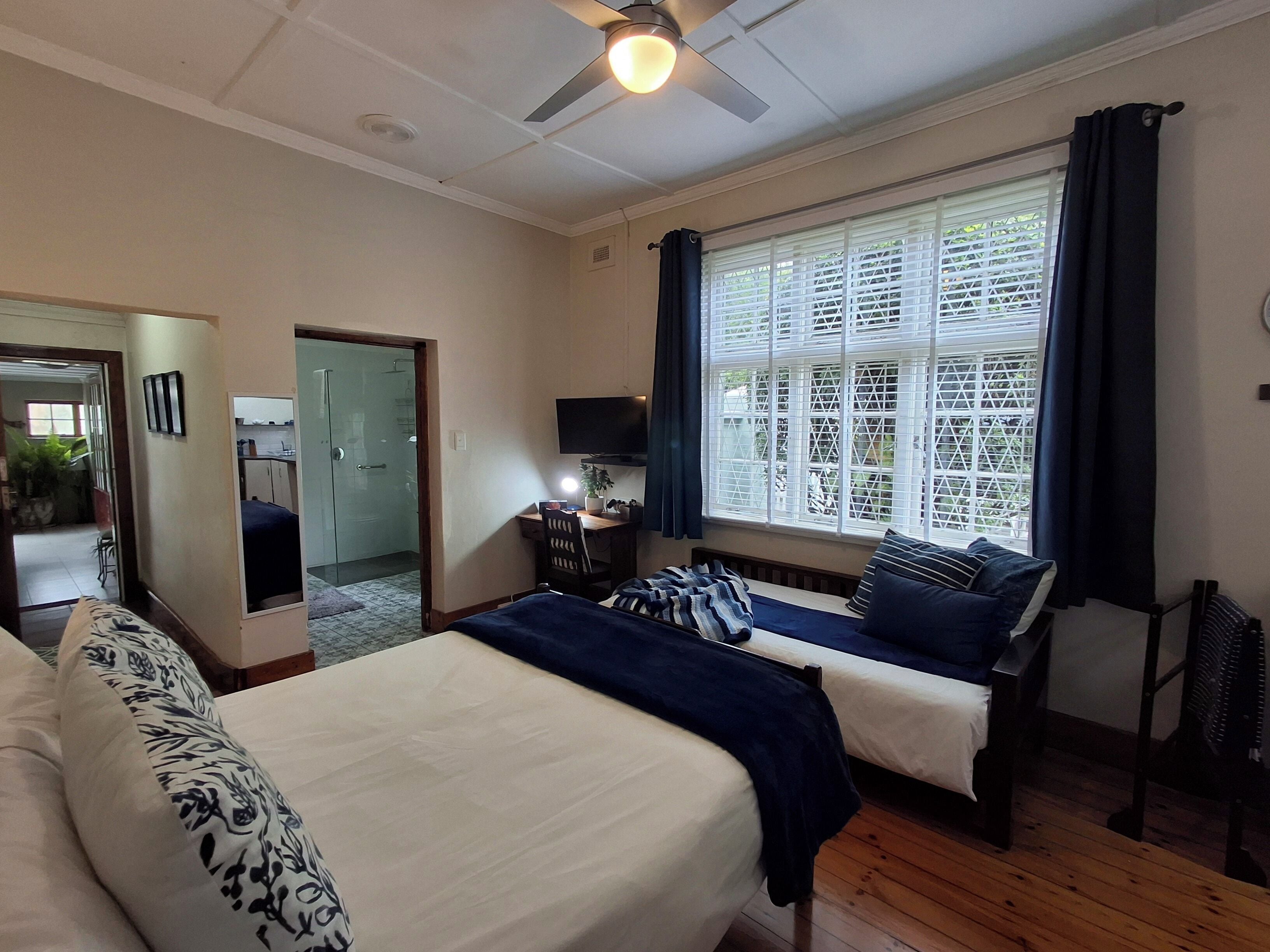 A bedroom with two beds, a desk, a TV, and a large window with blinds.