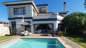 Una casa de dos pisos con piscina, un patio con sillones de descanso y una zona de estar techada al aire libre.