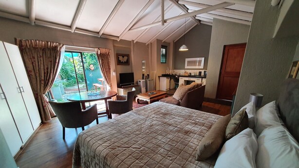 Un dormitorio con cama, escritorio y silla. Se ve una ventana con cortinas, un televisor y una kitchenette al fondo.