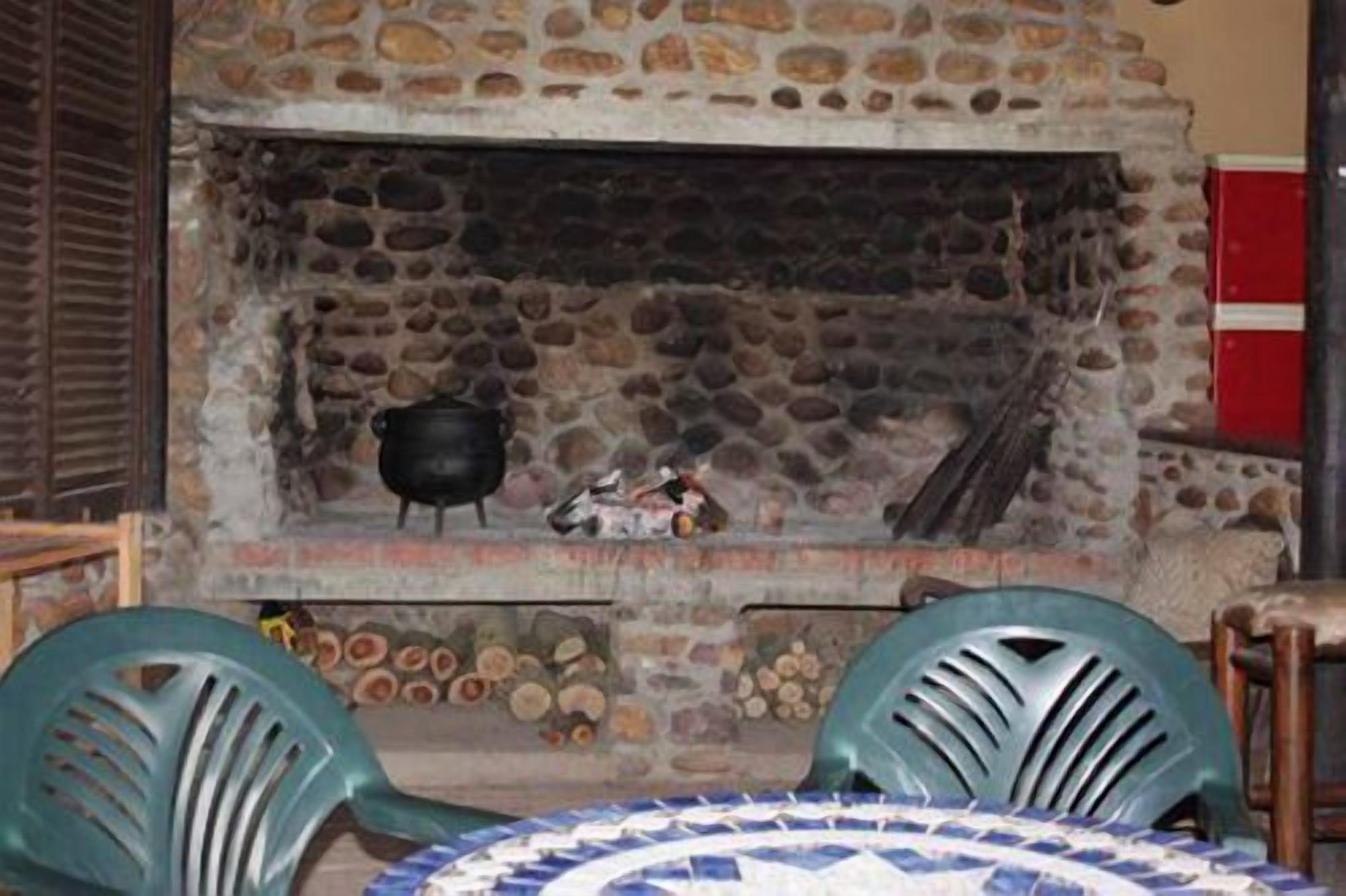 A stone fireplace with a pot on the hearth, logs stacked below, and a table with chairs in front.