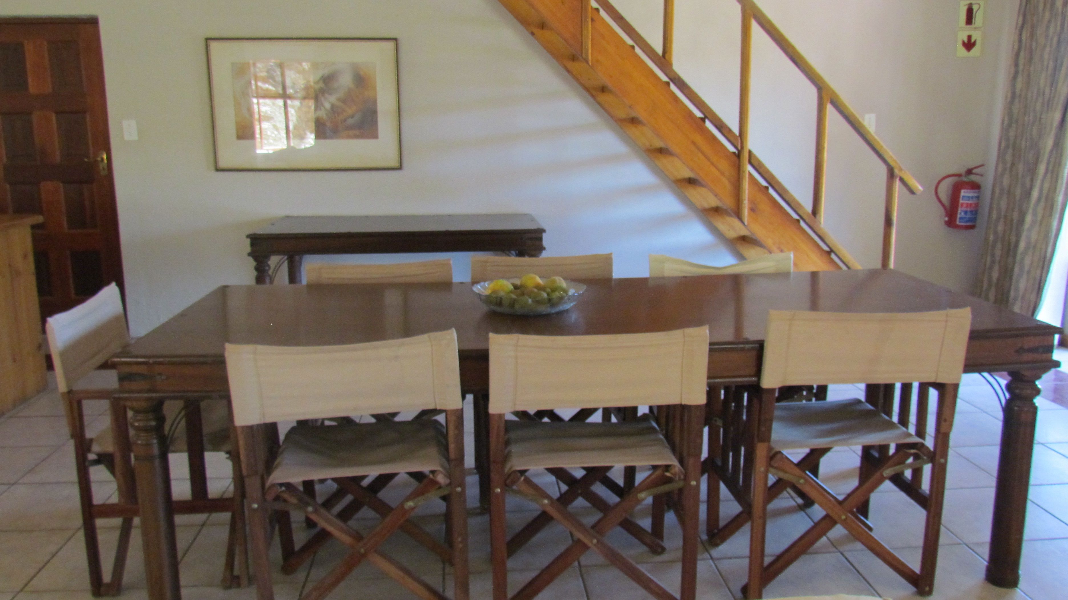 Un coin repas avec une table en bois, six chaises, un saladier de fruits, un escalier et un extincteur.