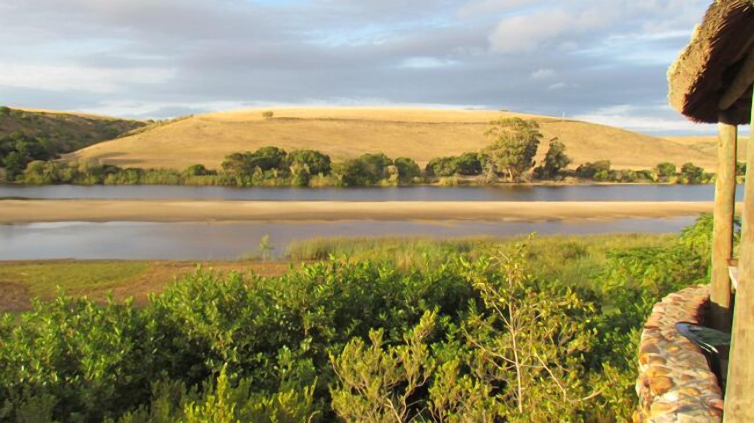Vista de un lago con una colina cubierta de hierba al fondo y una estructura con techo de paja en primer plano.