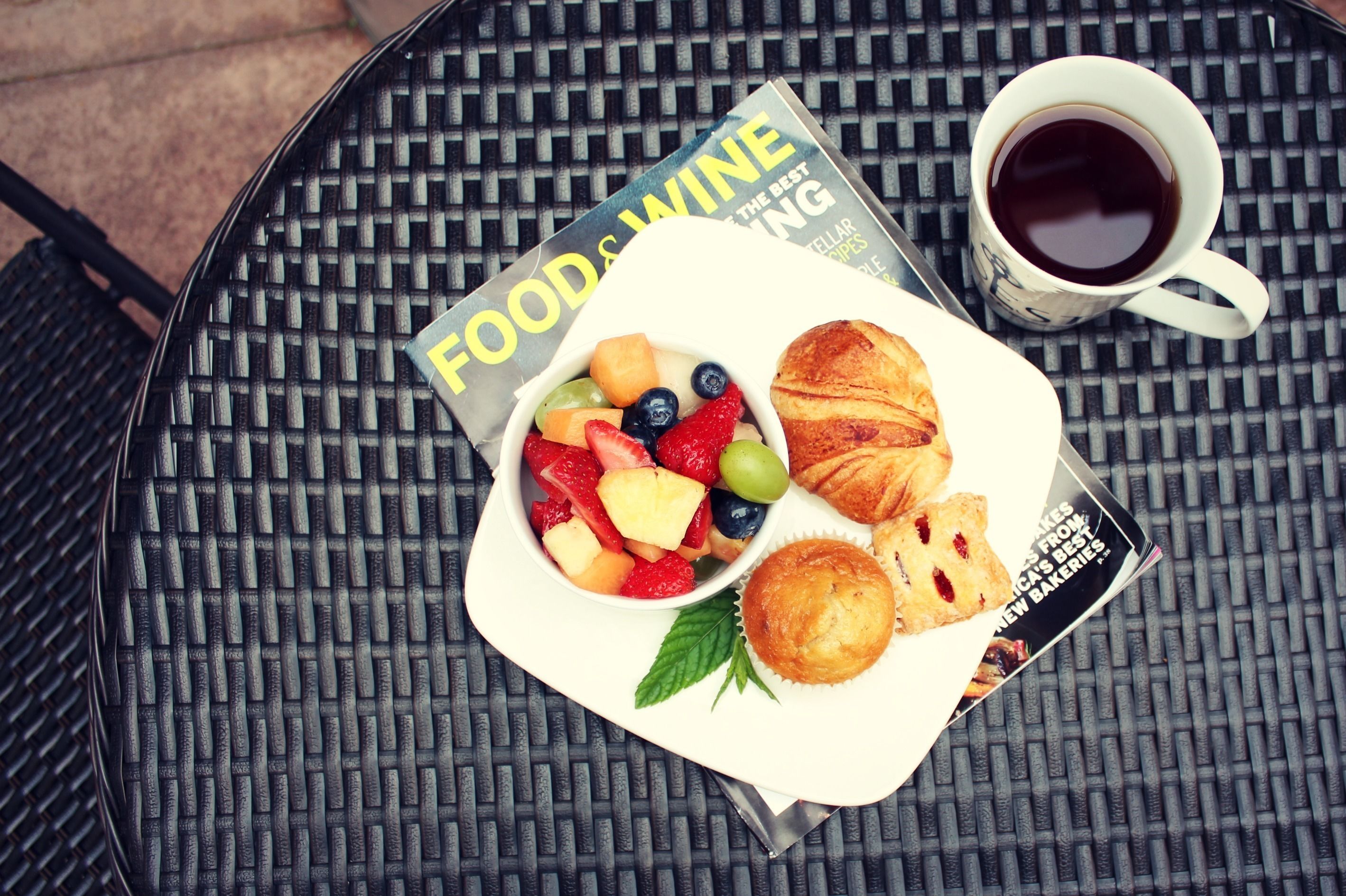 A table with a magazine, a cup of coffee, a plate of fruit, and pastries.