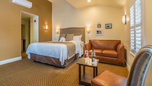 Une chambre d’hôtel avec un lit, un canapé en cuir, une petite table avec un verre à vin et une fenêtre avec des stores.
