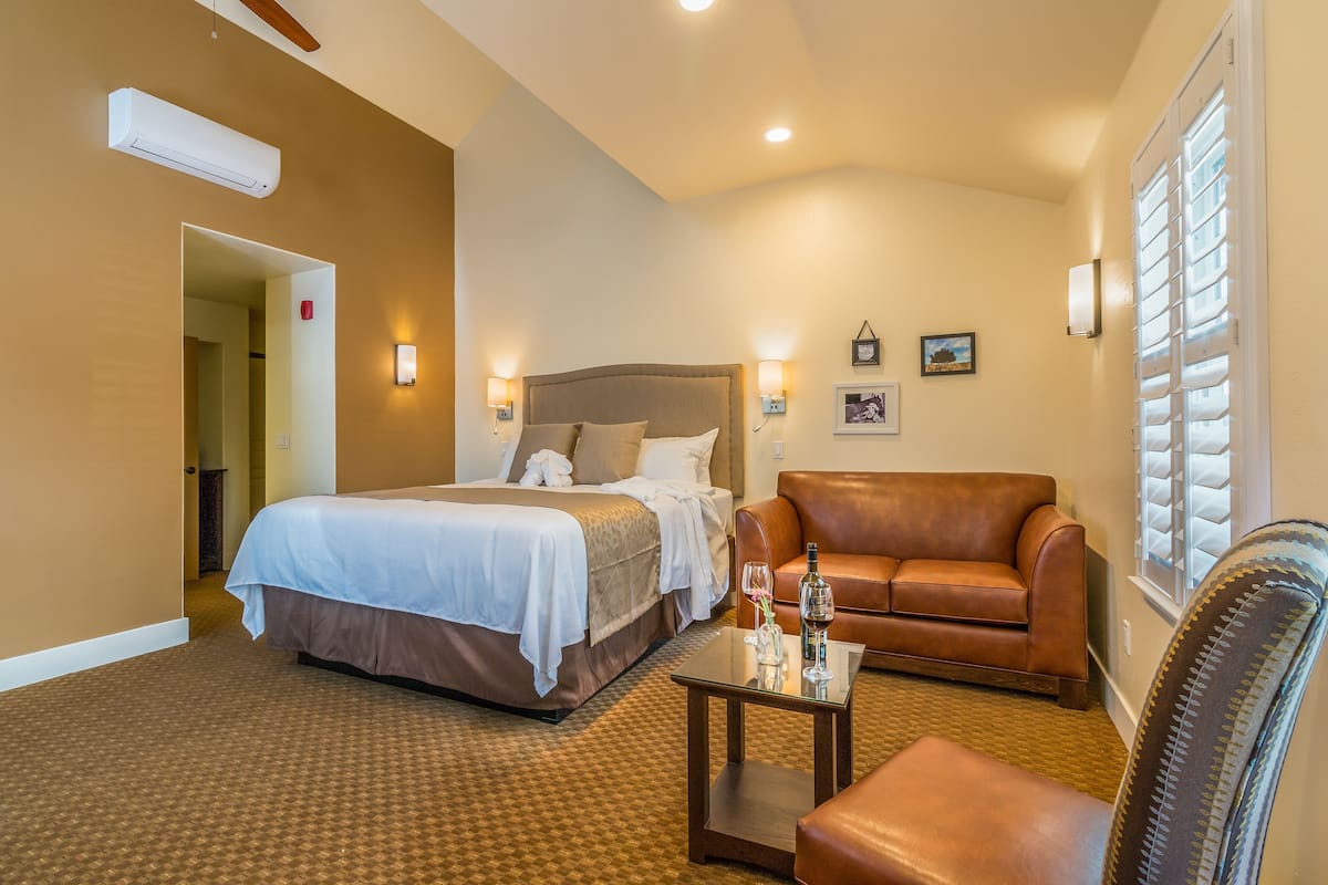 Habitación de hotel con cama, sofá de cuero, una mesita con una copa de vino y ventana con persianas.