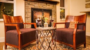 Un coin salon accueillant avec deux chaises en bois et une petite table ronde, une cheminée en briques et un vase décoratif contenant des verres à vin.