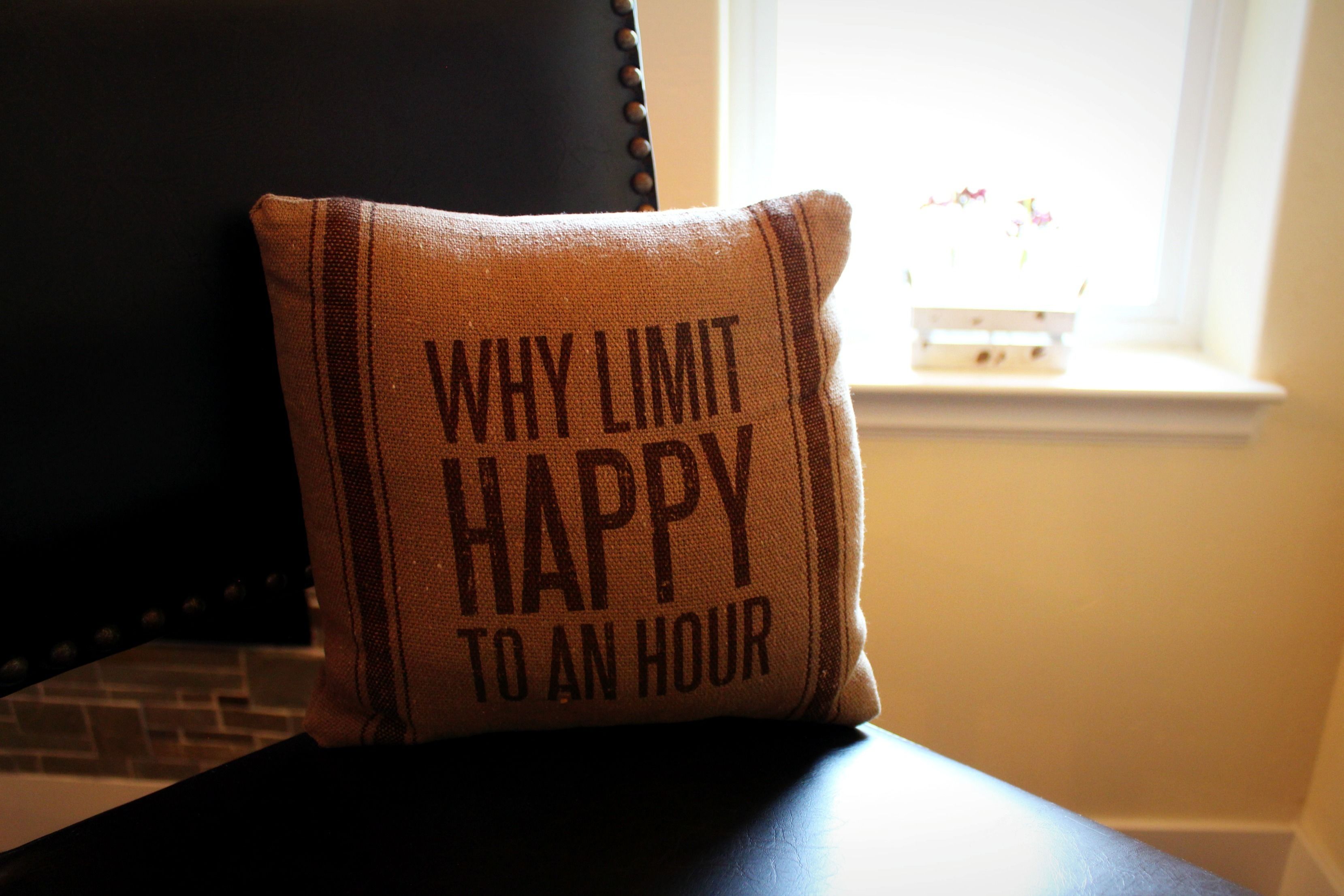 Un cojín con el texto "WHY LIMIT HAPPY TO AN HOUR" sobre una silla de color oscuro.