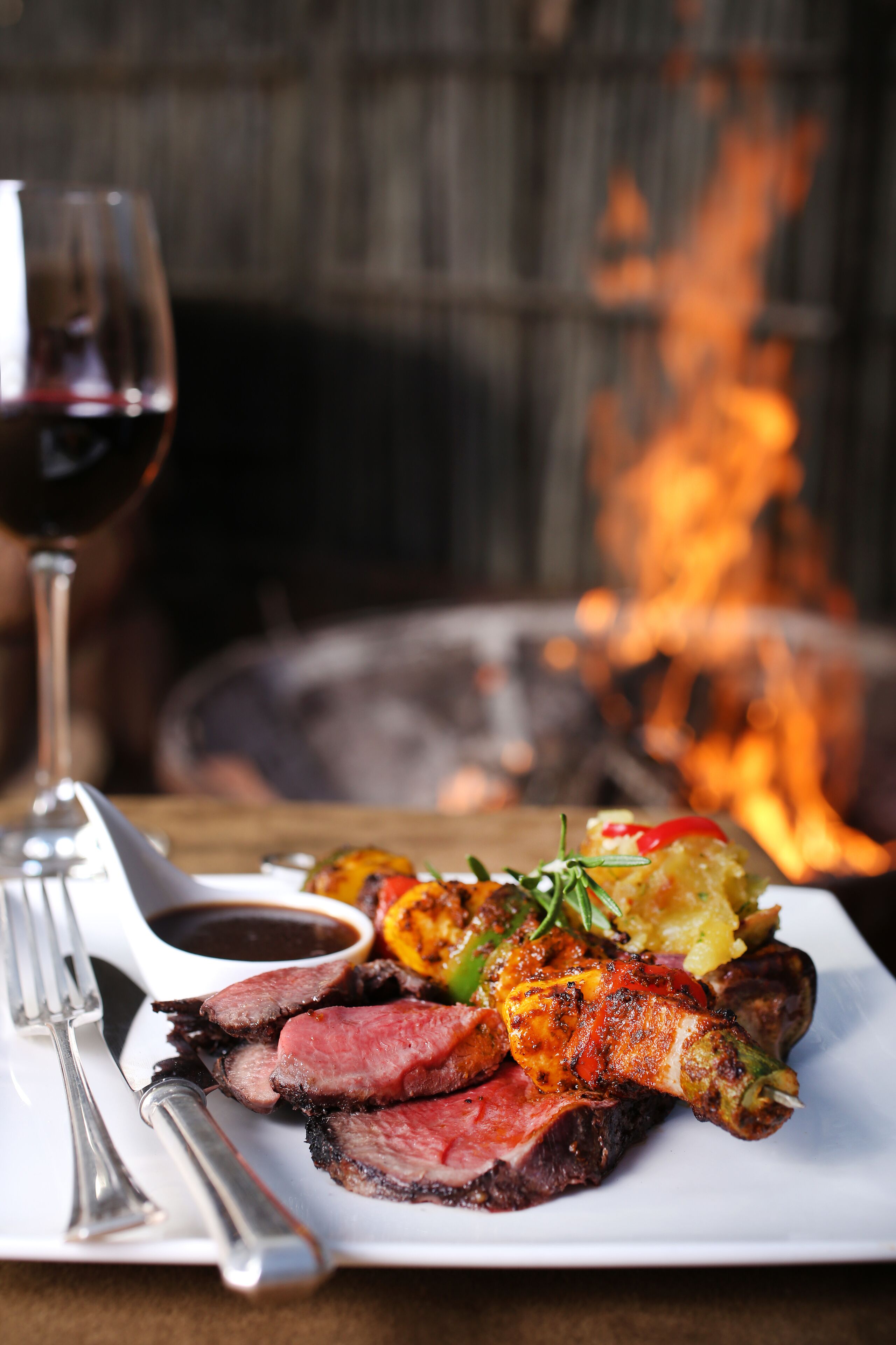 A plate of grilled meat with vegetables and a glass of red wine, set against a backdrop of a fire.