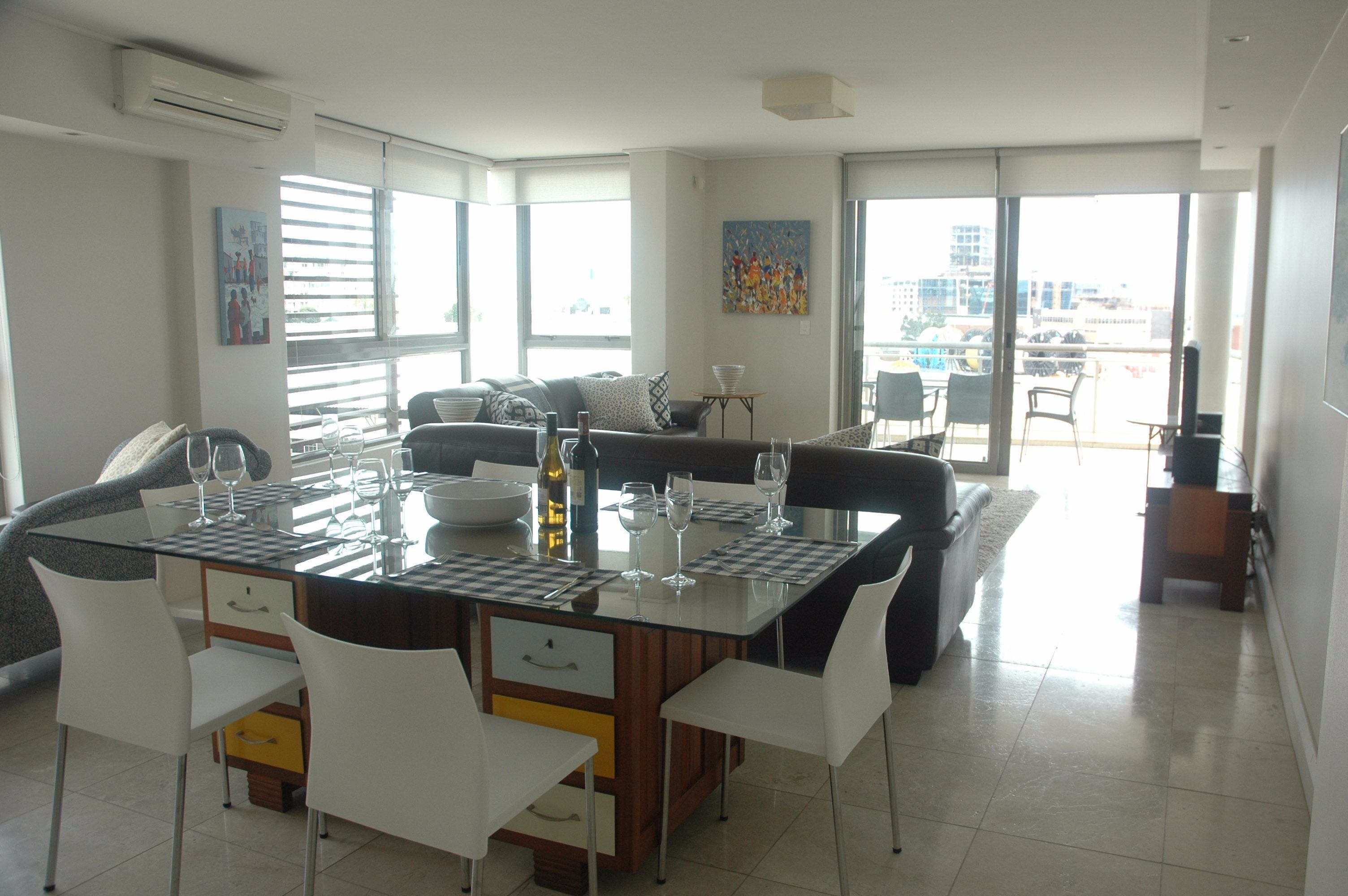 A modern dining area with a glass-top table, white chairs, and a central island with a bottle and glasses.