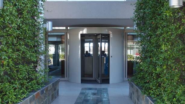 A modern building entrance with a glass door, surrounded by greenery and stone walls. - 501 Canal Quays (Cape Town)