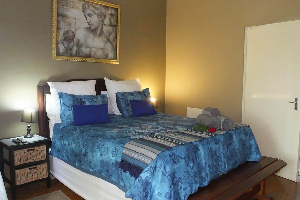 Un dormitorio con cama, almohadas, mesita de noche, lámpara, un cuadro y una puerta.