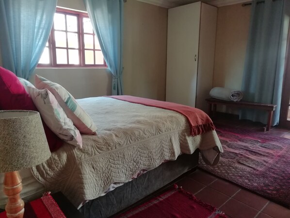 Un dormitorio con cama, almohadas, lámpara, armario y ventana con cortinas.