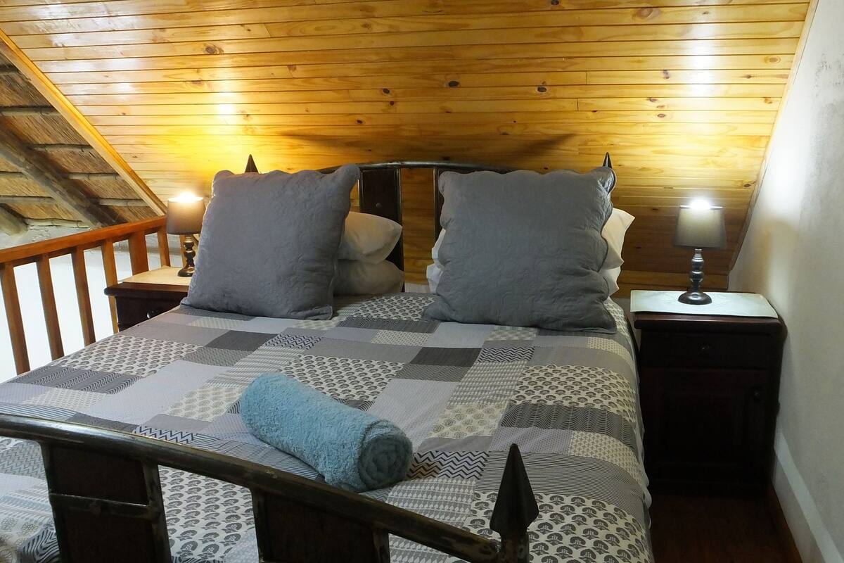 Un dormitorio con techo de madera, cama con ropa de cama estampada, dos almohadas, una lámpara de noche y cabecera de madera.