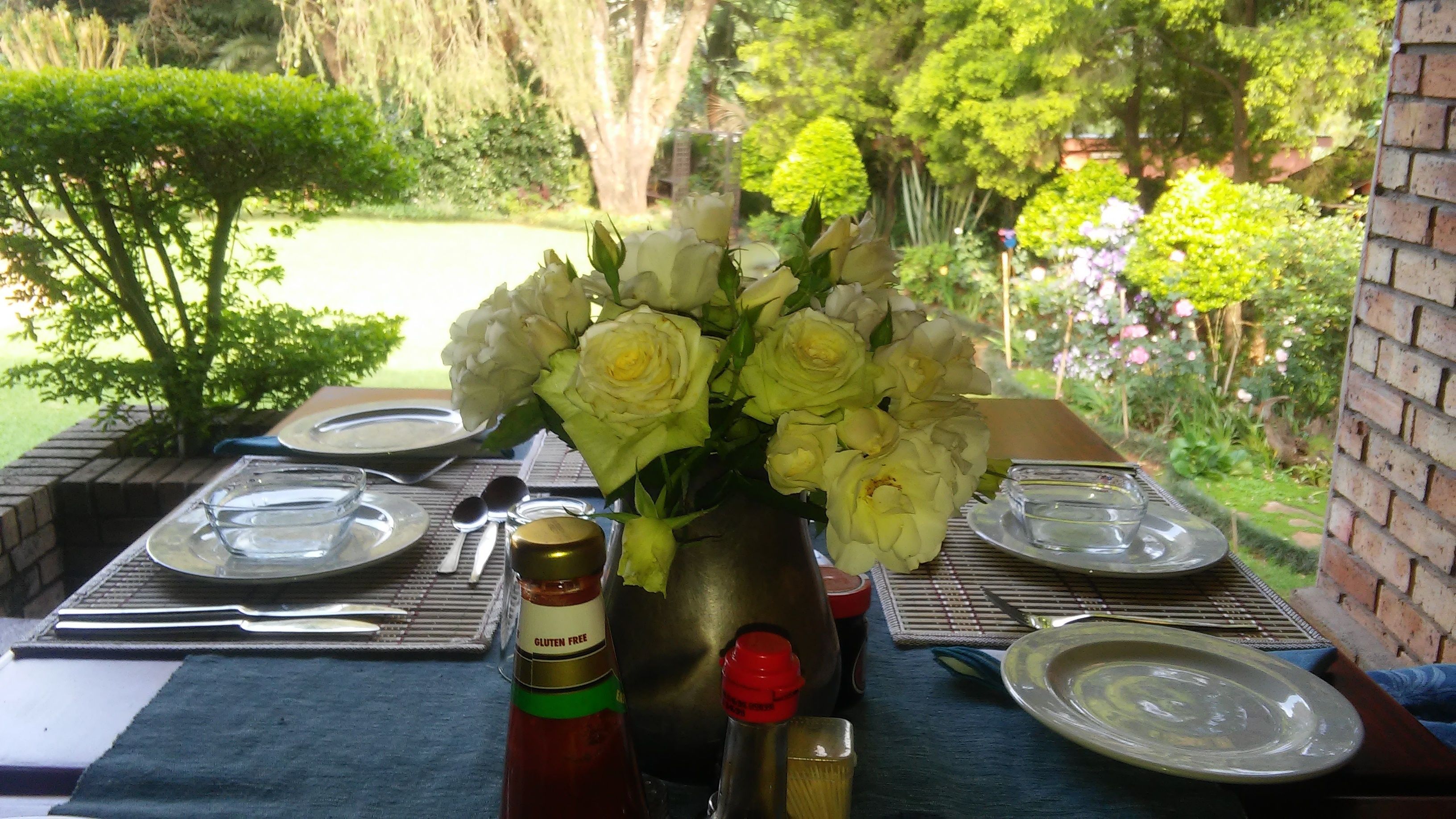 A table set outdoors with a vase of white flowers, plates, cutlery, and a bottle of gluten-free ketchup.
