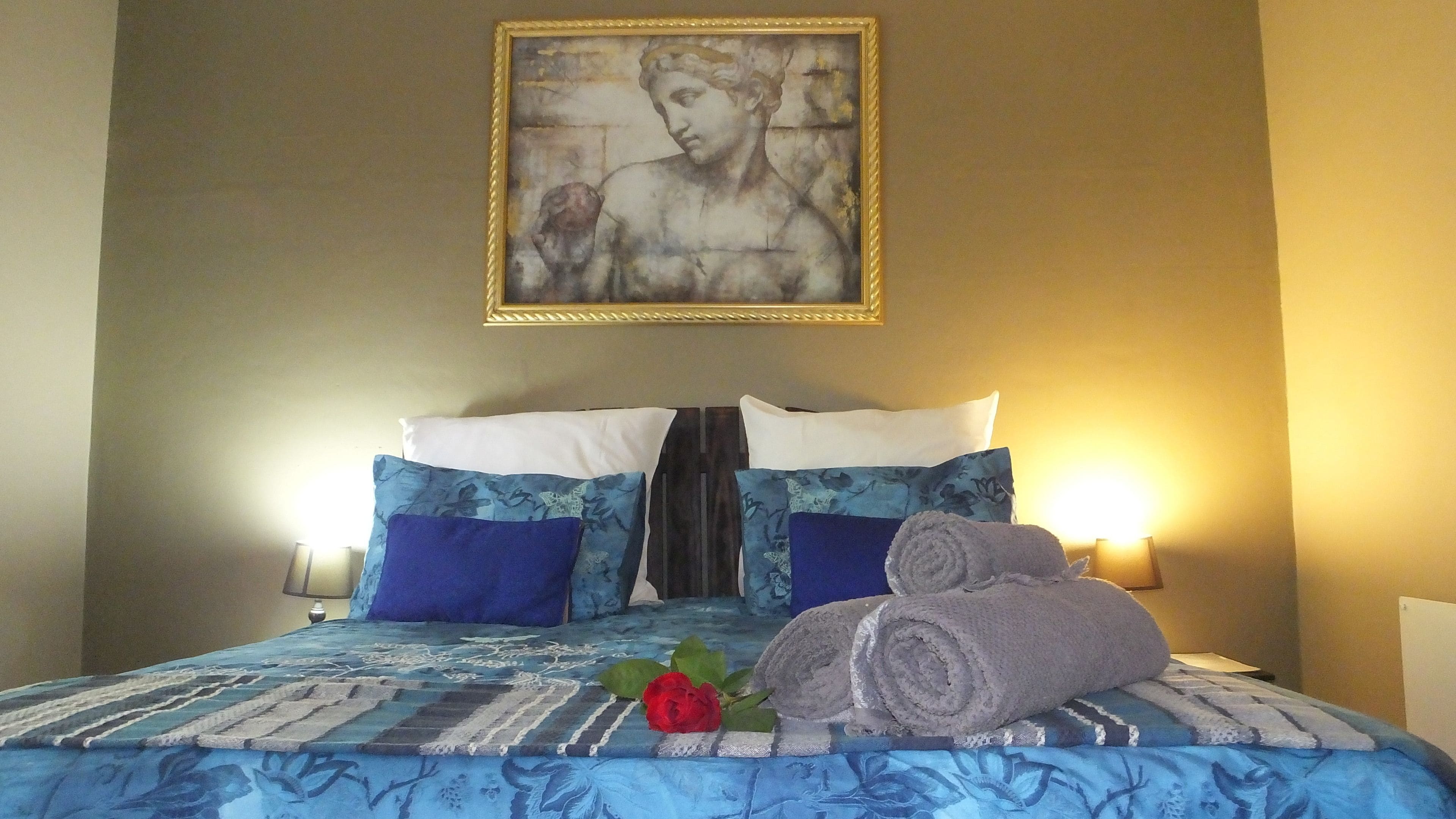 A neatly made bed with blue and white bedding, a framed artwork above, and a red rose on the bed.