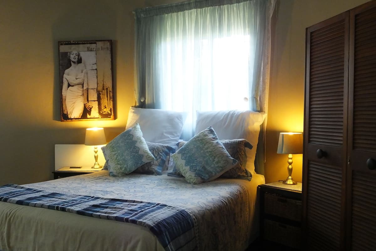 Una cama bien hecha con ropa de cama estampada, dos lámparas de noche, una mesita de noche, un armario y un cuadro en la pared.
