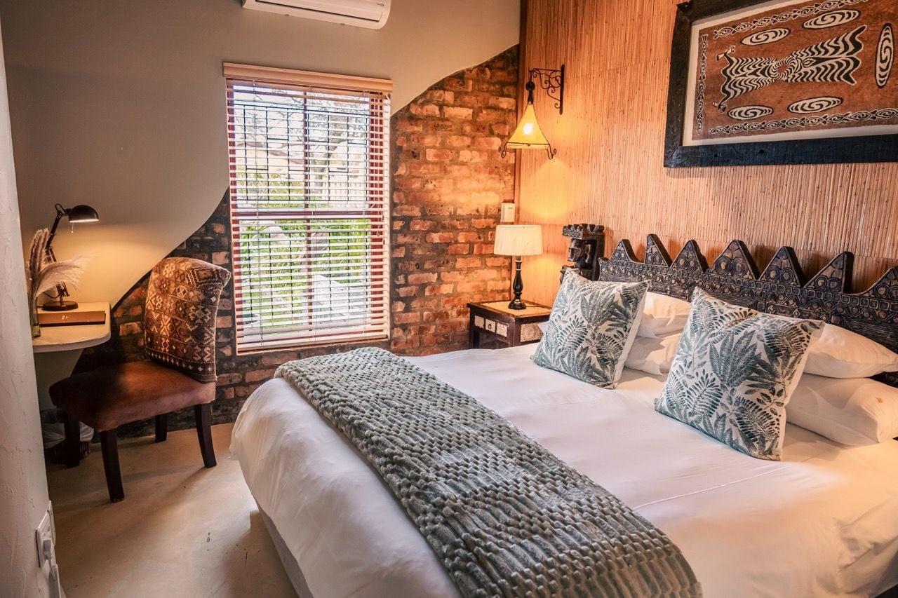 A bedroom with a brick wall, a bed with patterned pillows, a desk with a lamp, and a window with blinds.