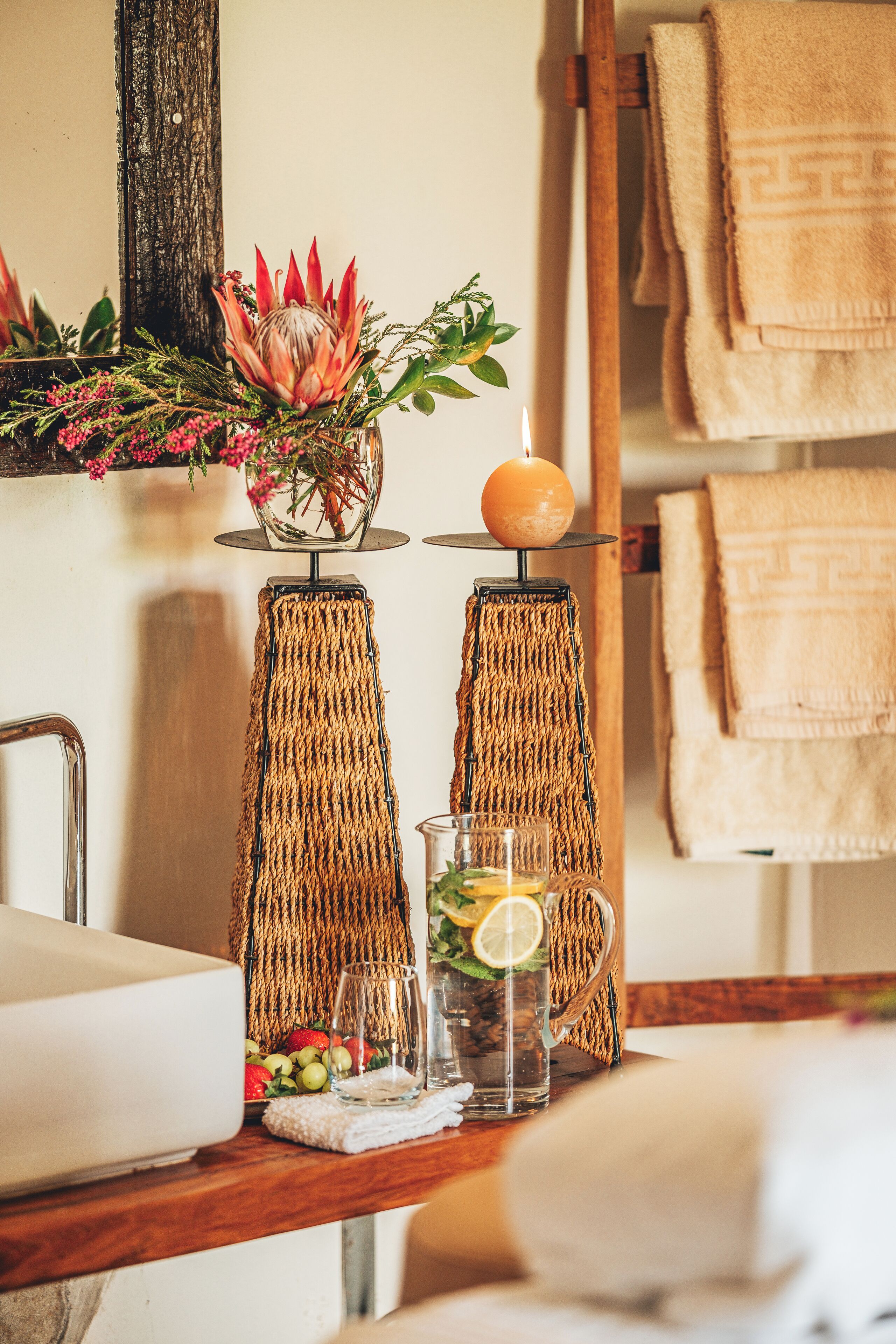 A bathroom with a wooden shelf holding a decorative plant, a glass of water with lemon, and a towel rack with folded towels.