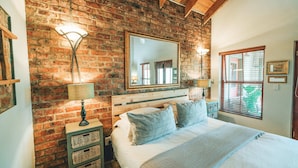 A bedroom with a brick wall, a large mirror, two bedside lamps, a bed with a patterned throw, and a window with blinds. - Villa Chad Luxury Guesthouse (Kleinmond)
