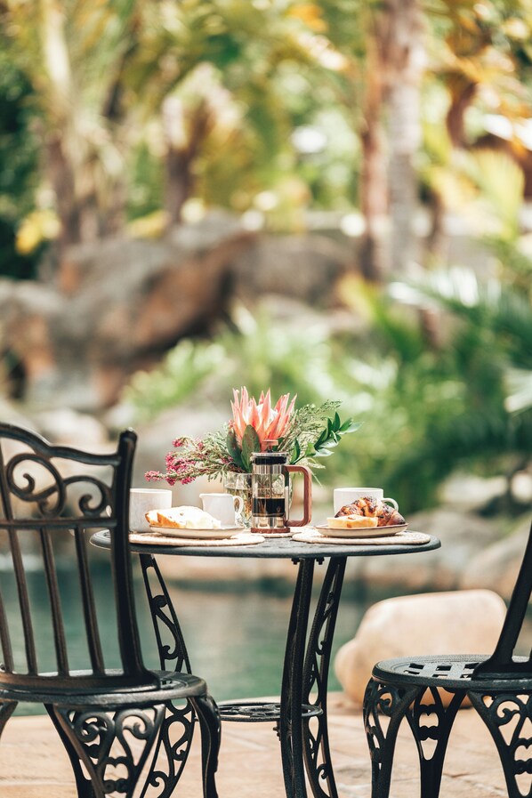 A table set with a coffee pot, cups, and pastries, surrounded by chairs, with a natural backdrop.