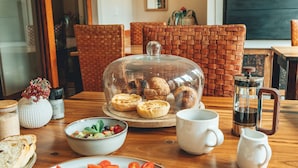 A breakfast setting with a wooden table, a glass dome displaying pastries, a bowl of fruit, and a plate with eggs. - Villa Chad Luxury Guesthouse (Kleinmond)