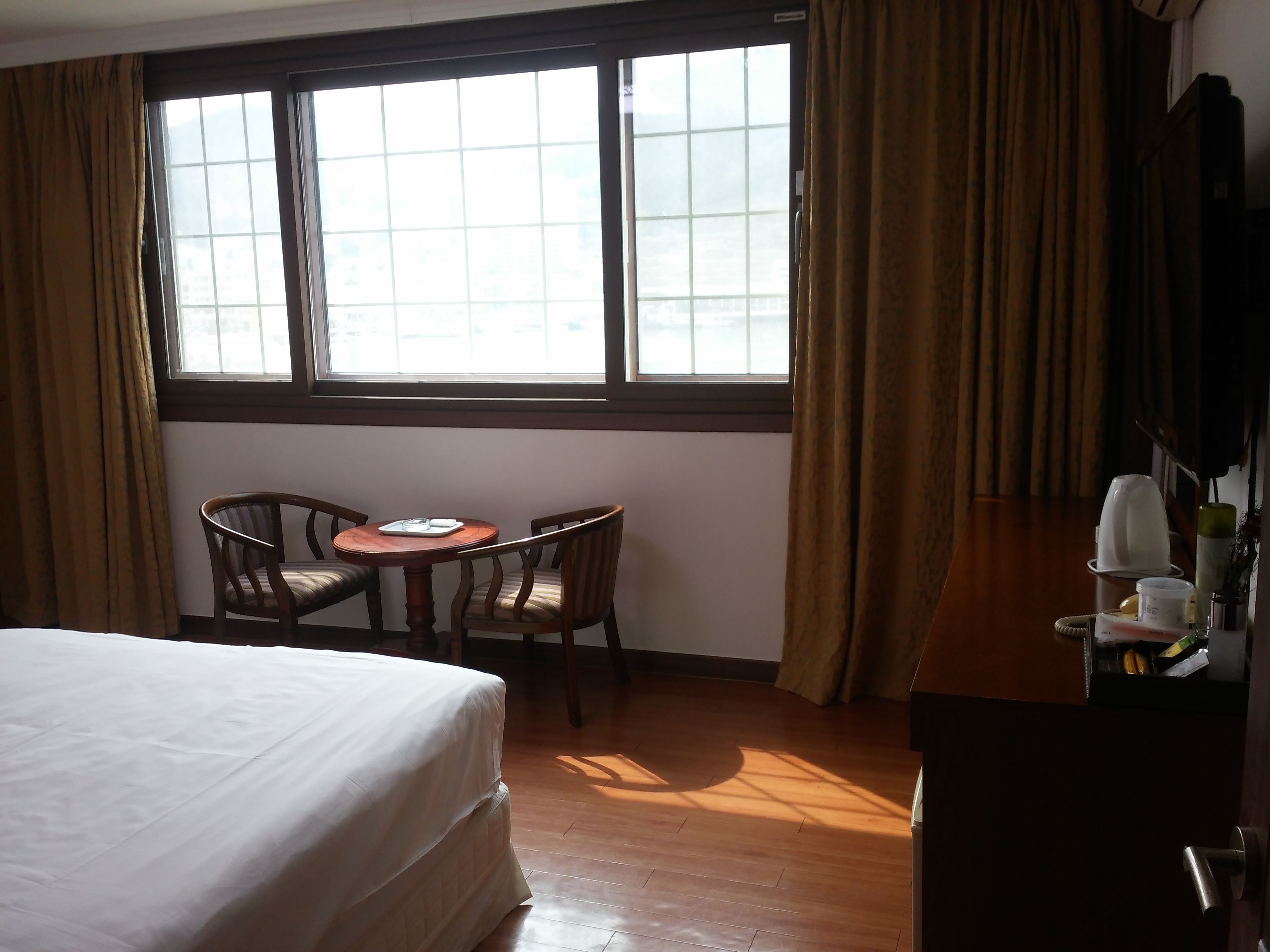Una habitación de hotel con una cama, una mesita redonda pequeña con sillas, una ventana con cortinas y un aparador de madera.
