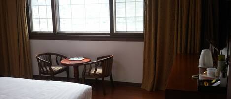 Una habitación de hotel con una cama, una mesita redonda pequeña con sillas, una ventana con cortinas y un aparador de madera.