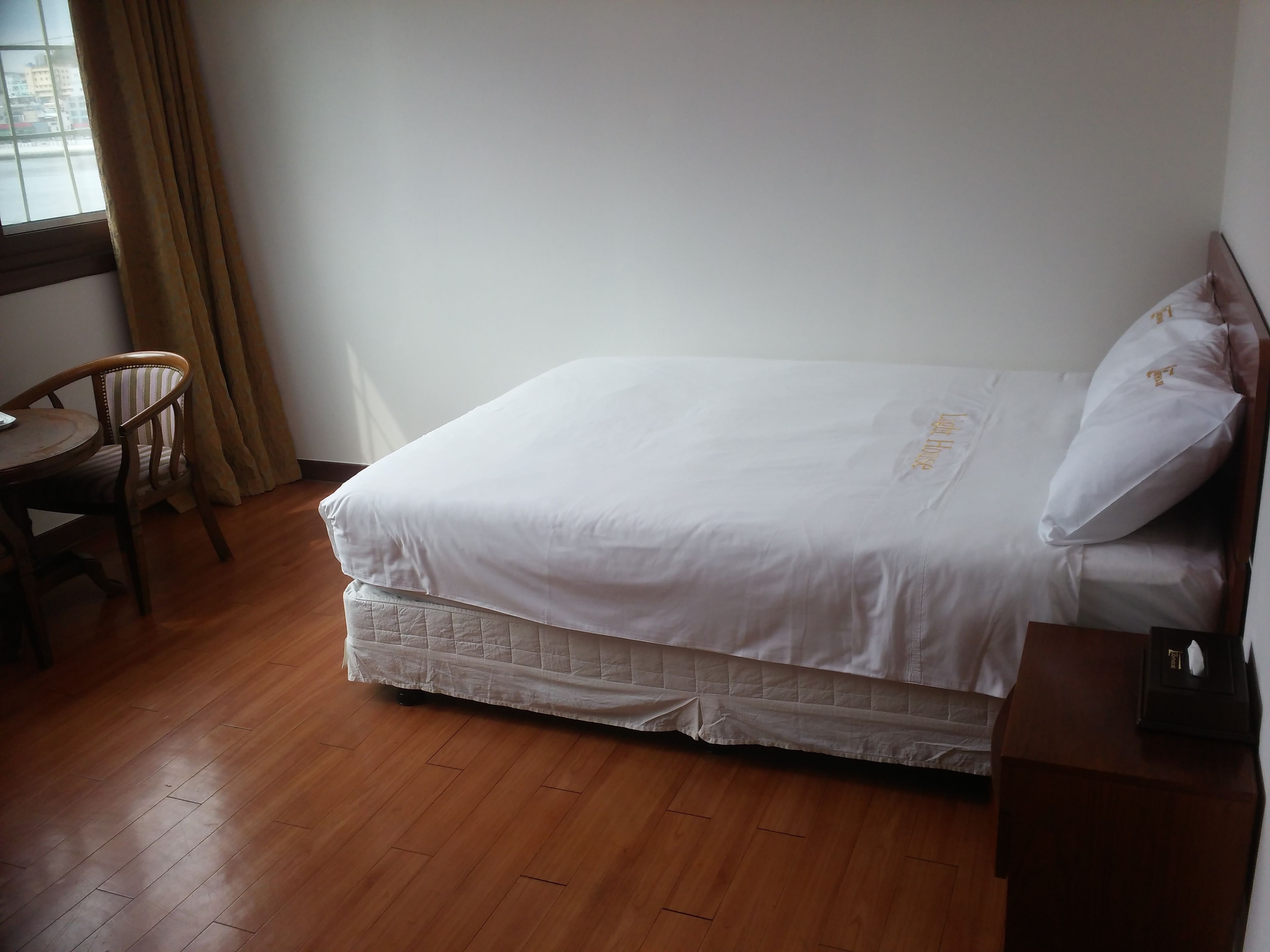 Una cama bien hecha con ropa blanca y un cubrecolchón acolchado, una silla de madera y una mesita con una caja de pañuelos encima.