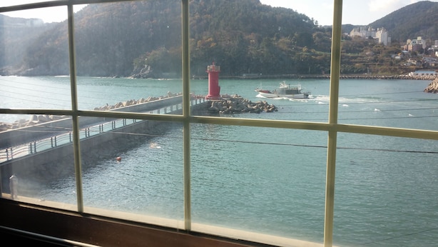 Vista a través de una ventana de una zona costera con un faro, barcos y una costa rocosa.