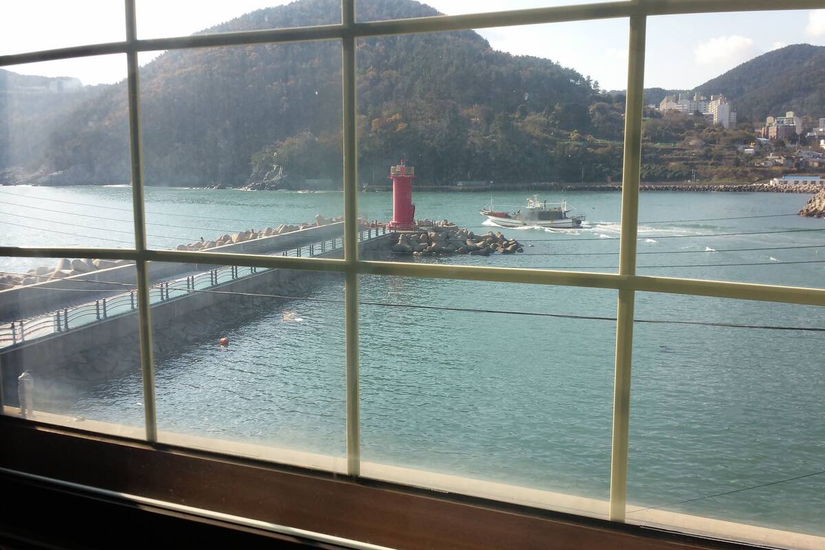 A view through a window of a coastal area with a lighthouse, boats, and a rocky shoreline.