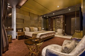 Una habitación de hotel moderna con una cama grande, una zona de estar con una mesa de cristal y un rincón de descanso con una silla acolchada.