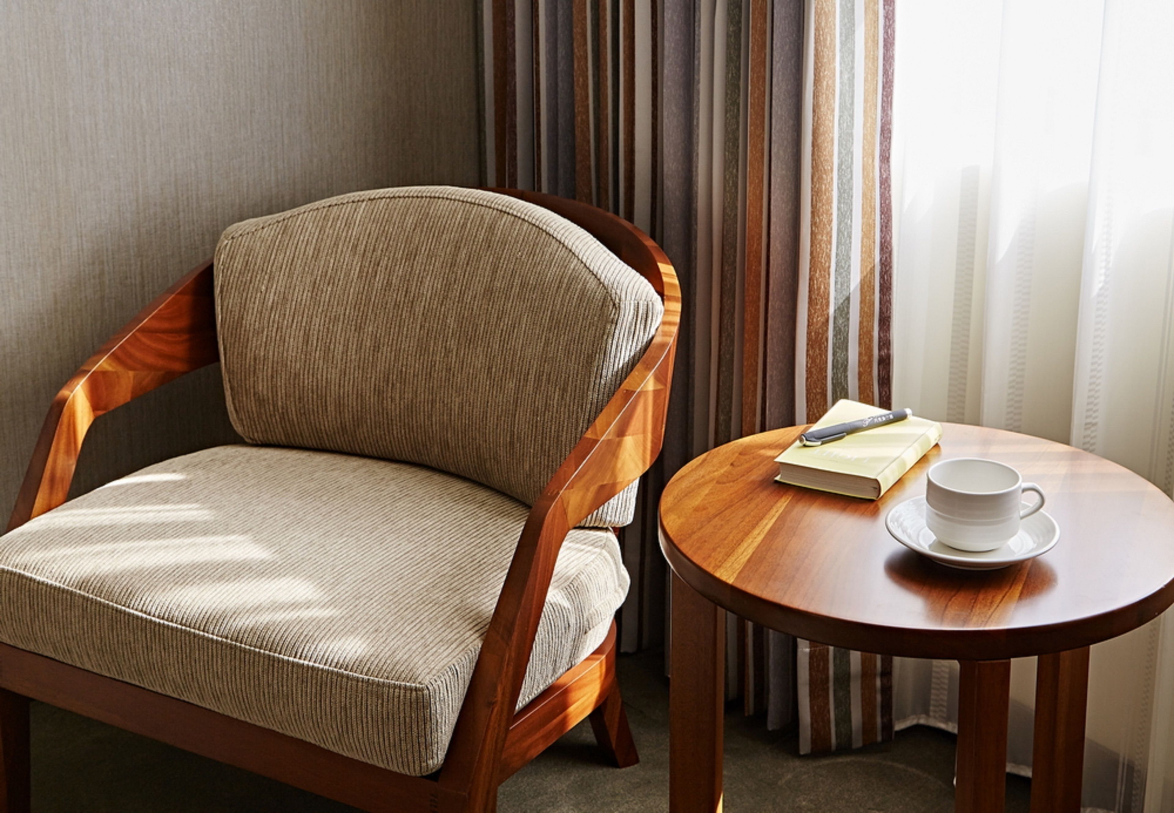 Un sillón de madera con asiento y respaldo acolchados, una mesita de madera con una taza y un libro, y una ventana con cortinas.