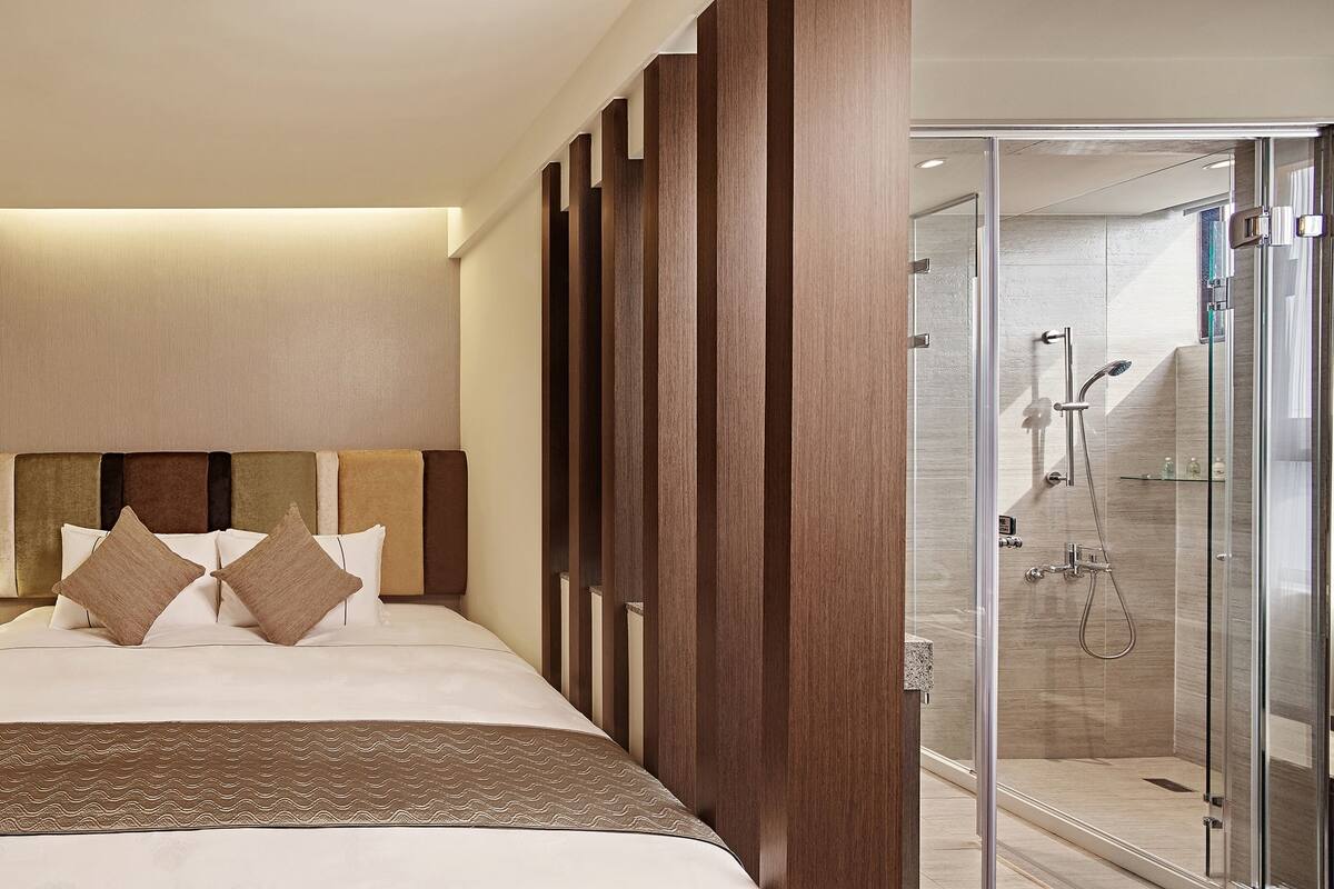 A hotel room with a bed, a nightstand with a lamp, a telephone, and a shower area with glass doors.