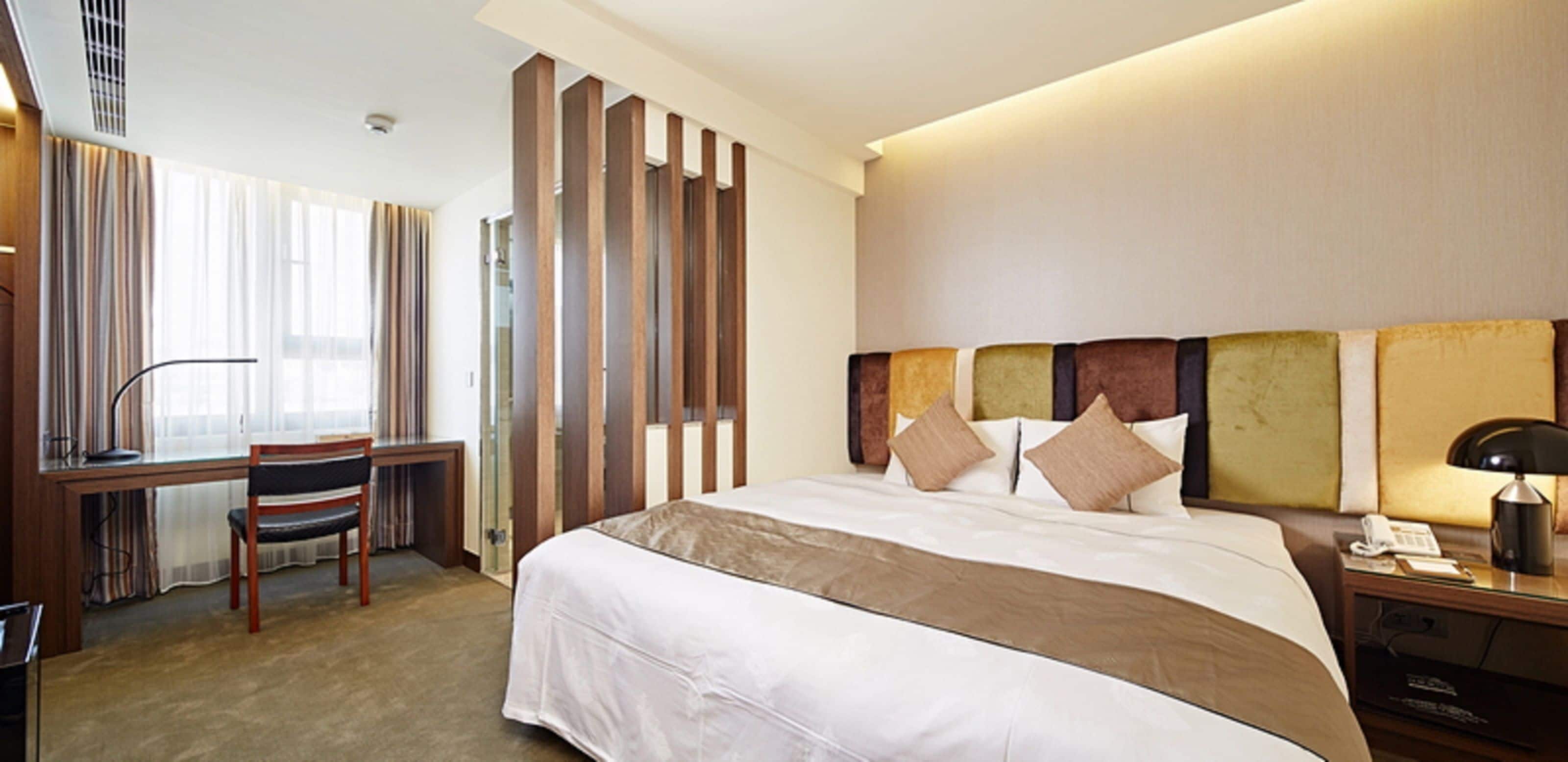 A hotel room with a large bed, a desk, a chair, and a nightstand with a lamp.