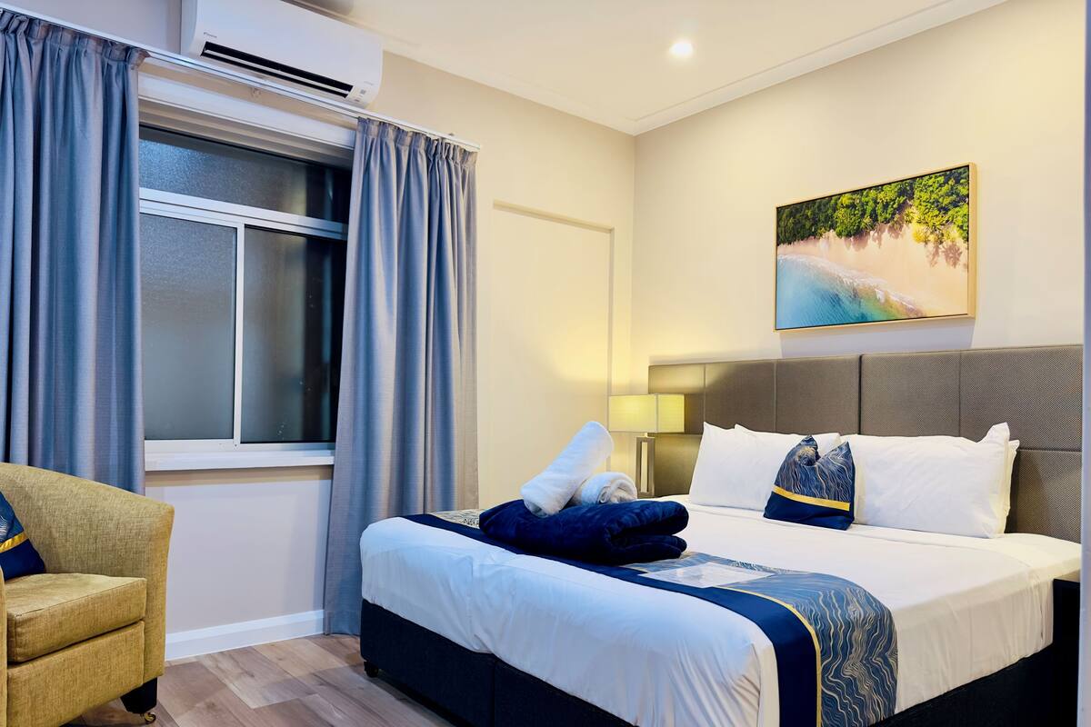 Une chambre d’hôtel avec un lit, une chaise, une fenêtre avec des rideaux, un tableau au mur et une lampe.