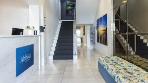 Zona de recepción de hotel con una escalera que conduce a los pisos superiores, un mostrador de recepción y una sala de espera con un sofá estampado con motivos florales.