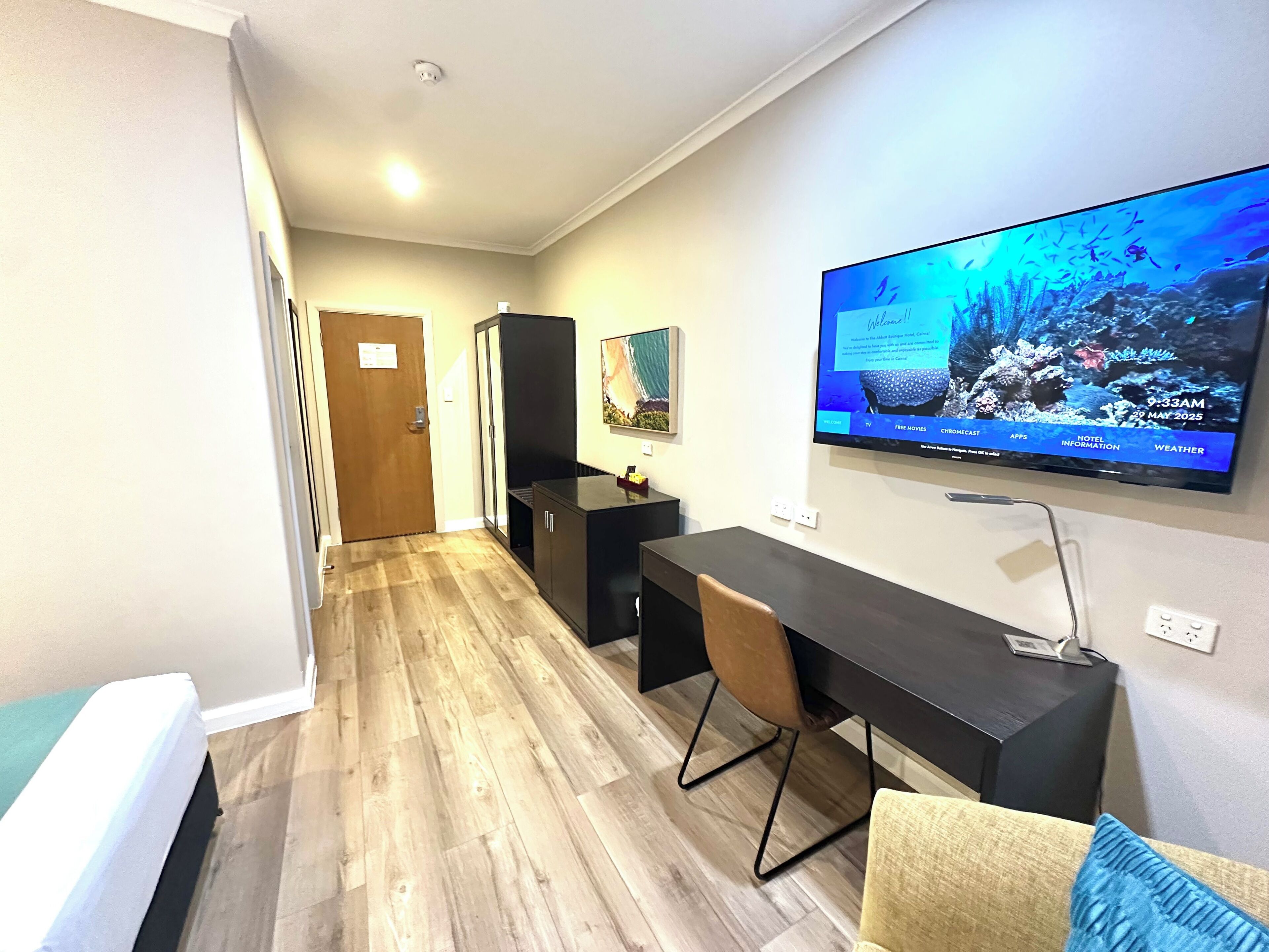 Habitación de hotel con piso de madera, televisor montado en la pared mostrando un mensaje de bienvenida, escritorio con silla y una puerta.