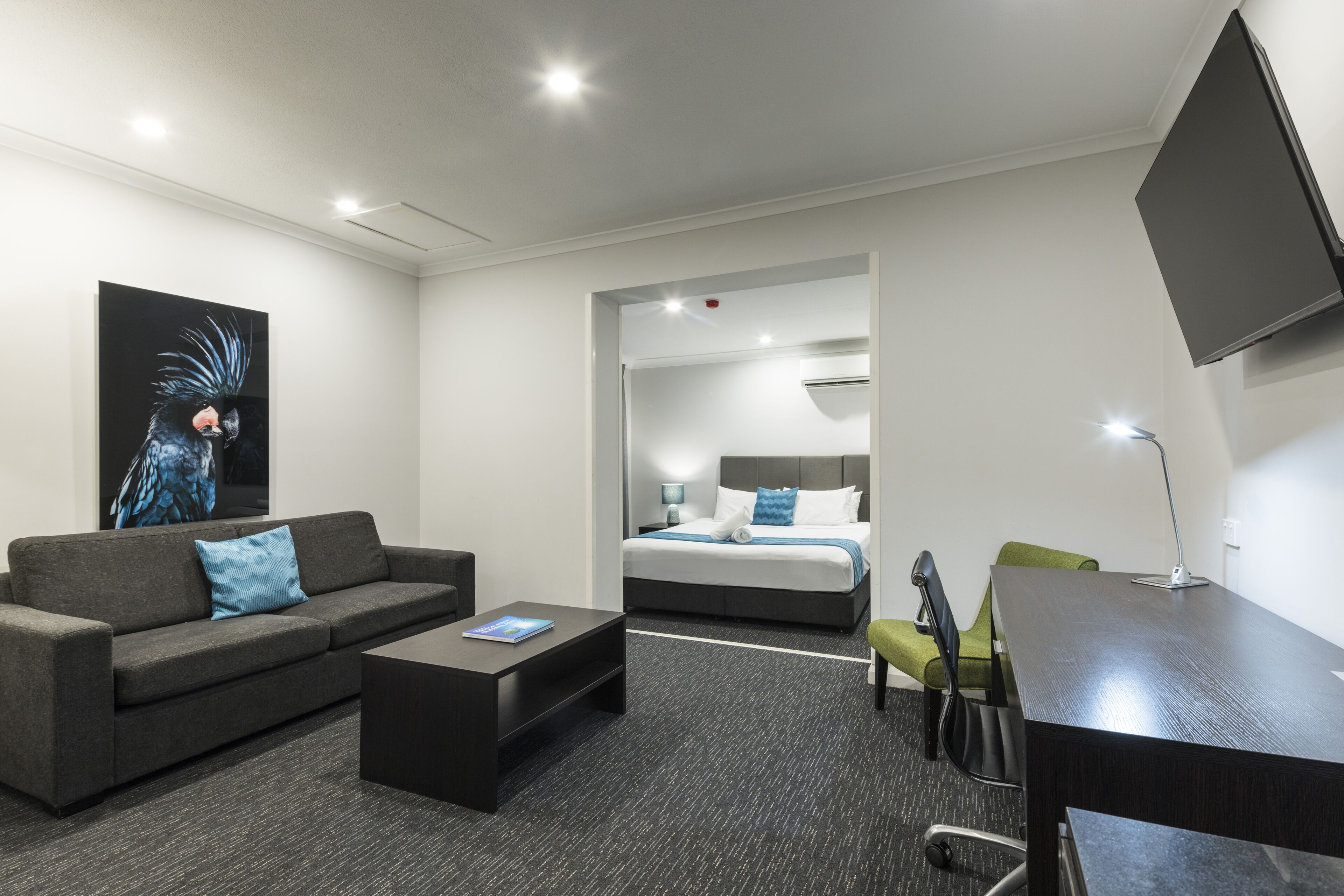 A modern hotel room with a gray sofa, a small coffee table, a desk, and a bed with a blue and white bedspread.