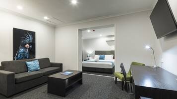 A modern hotel room with a gray sofa, a small coffee table, a desk, and a bed with a blue and white bedspread.