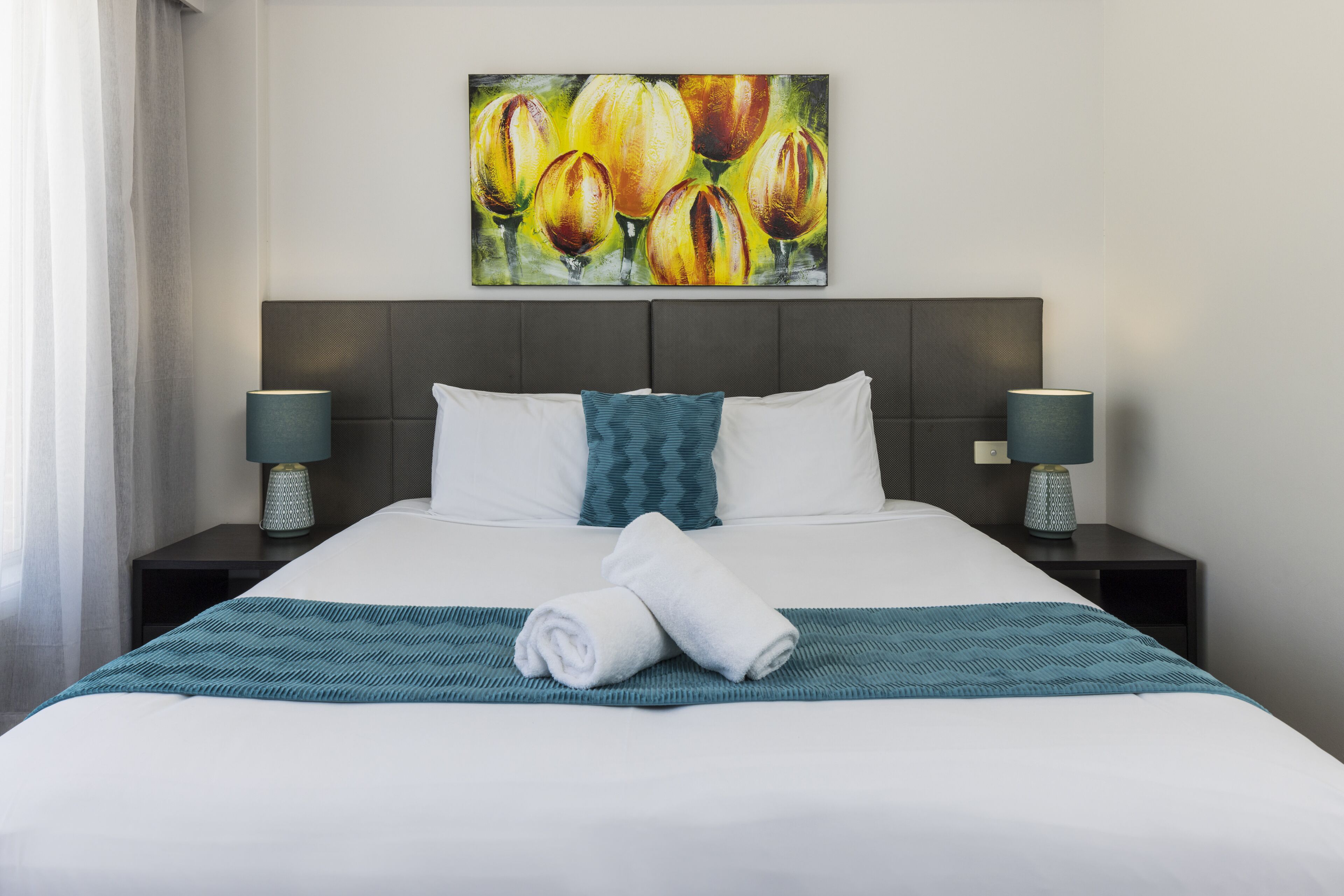 Una cama bien hecha con una manta color turquesa y toallas enrolladas, un cabecero oscuro y dos lámparas de noche a cada lado.