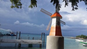 Un moulin à vent avec un corps rouge et blanc, une base en béton et une structure métallique. En arrière-plan, on aperçoit une étendue d’eau avec des bateaux et une côte lointaine.