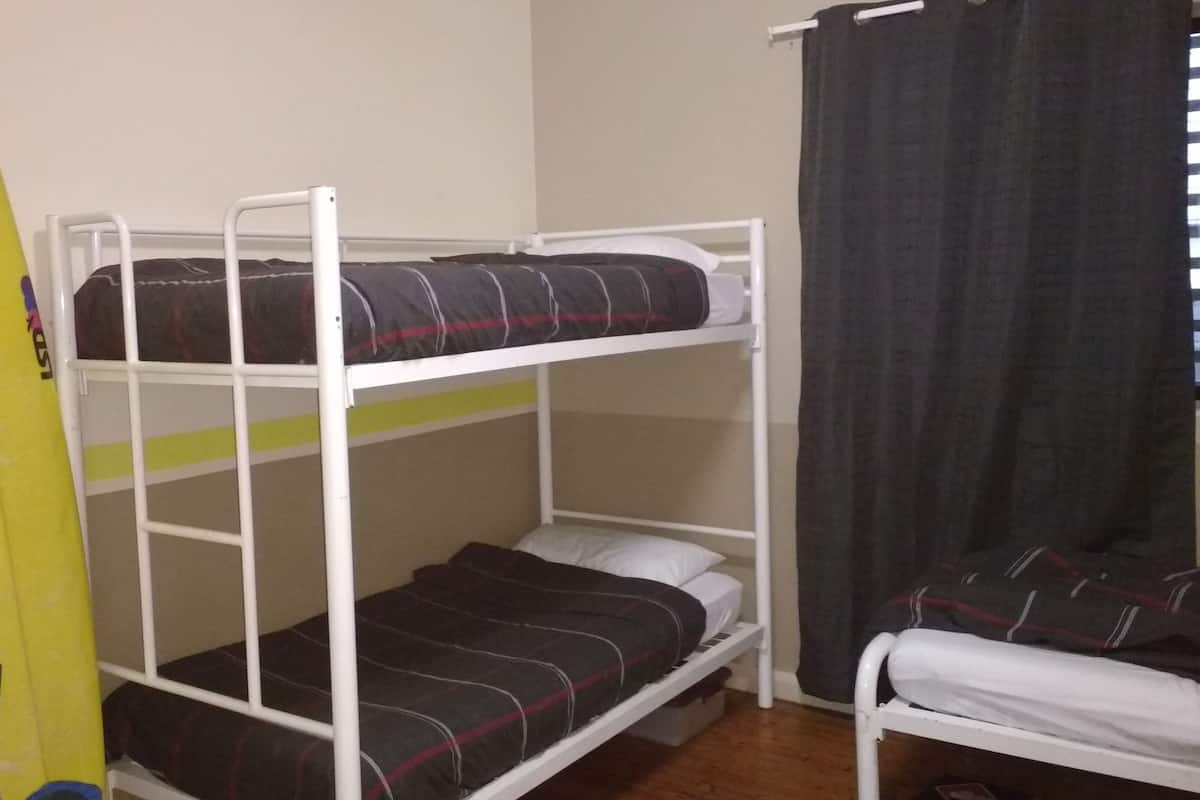 A bunk bed room with a surfboard leaning against the wall, a window with a curtain, and a small chair.