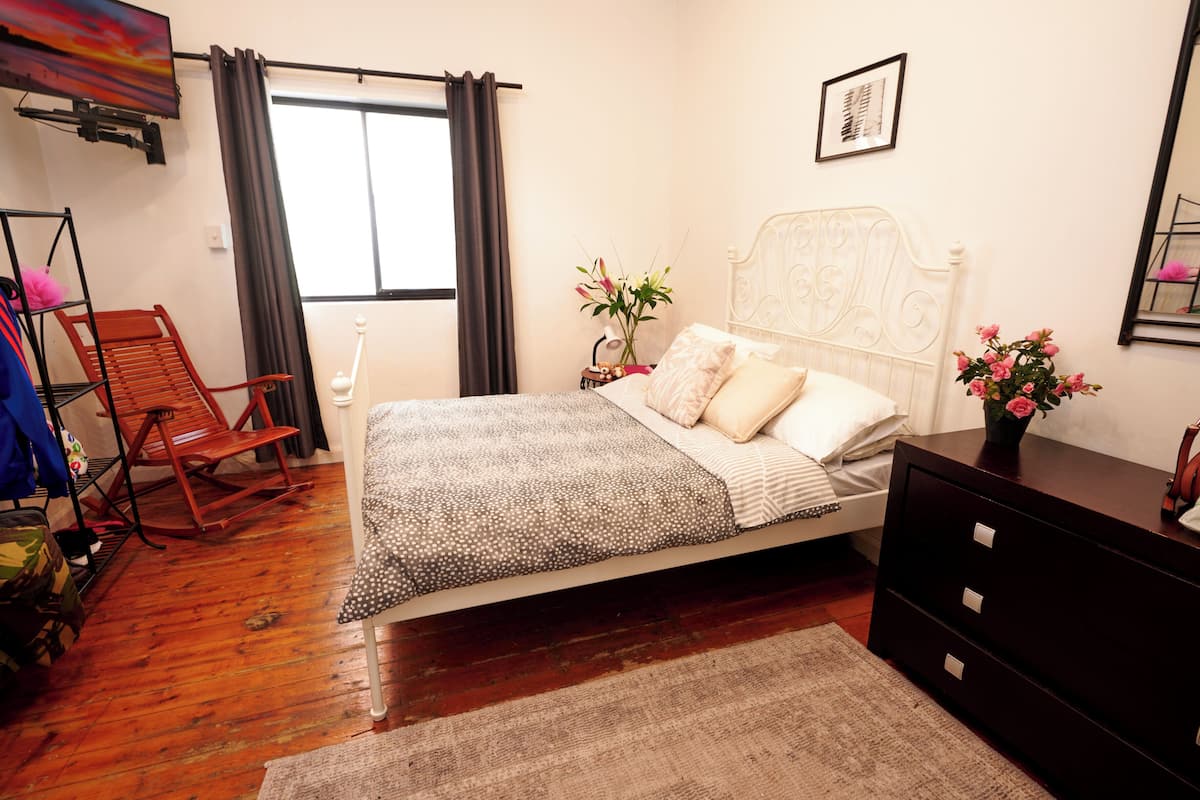 A bedroom with a bed, a dresser, a chair, a window with curtains, and a TV on the wall.